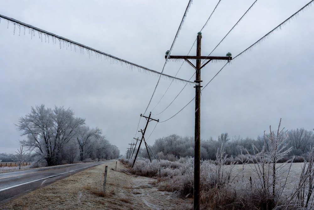 A line of wooden utility poles leaning over at increasing angles beside a rural road after a freezing-rain storm, the ice-coated conductors between them hanging in deep slack loops