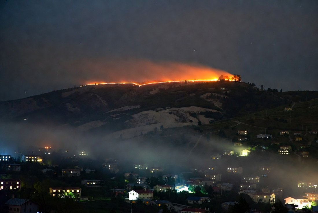 A fire line burning along a distant ridge at night above a valley of houses with their lights on, smoke drifting across the hillside