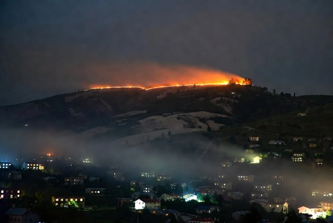 A fire line burning along a distant ridge at night above a valley of houses with their lights on, smoke drifting across the hillside