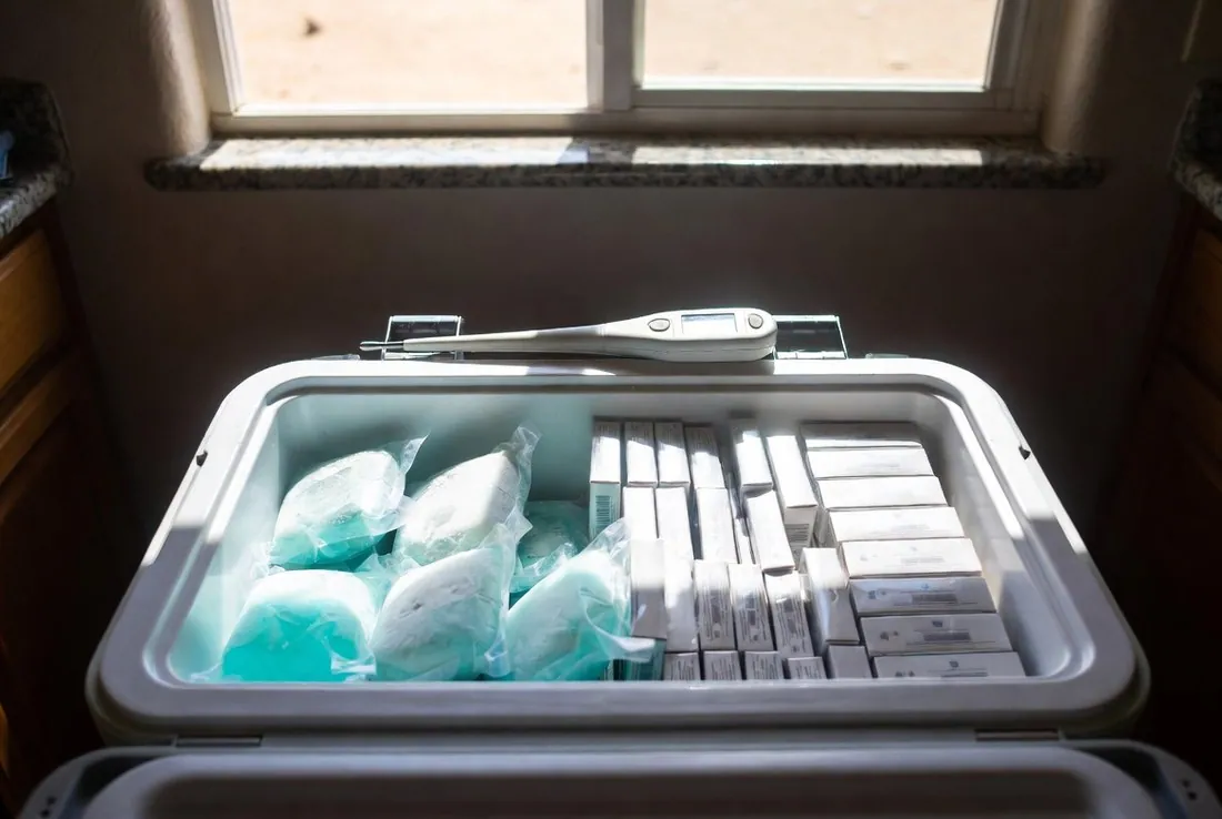 A cooler standing open on a kitchen counter packed with frozen gel packs on one side and rows of small closed medicine cartons on the other, a thermometer probe resting across the top