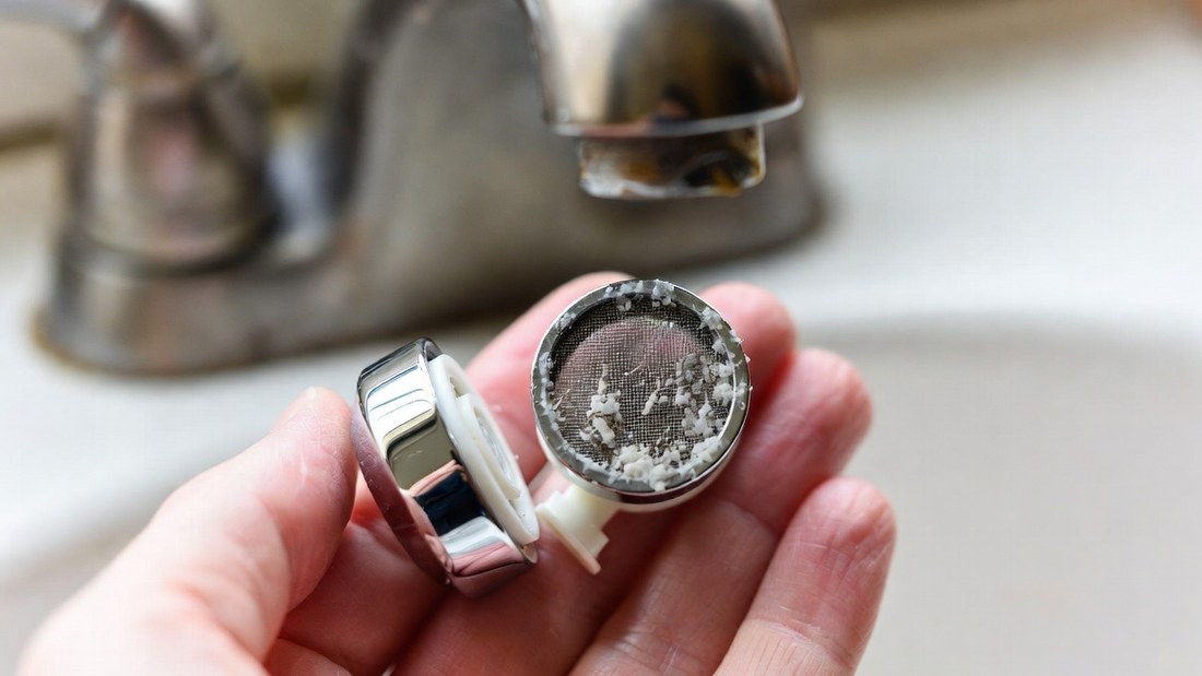 A faucet aerator unscrewed and held in the palm of a hand, its fine wire screen visibly packed with white lime scale and grit, with the bare threaded spout above it