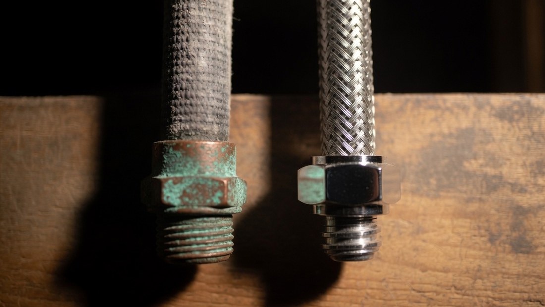 An old grey cloth-jacketed rubber supply line with a corroded green fitting standing beside a new braided stainless steel line with a clean chrome nut