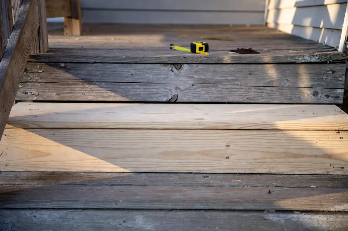 A set of grey weathered wooden porch steps with one fresh new board fitted into the middle tread, a tape measure and a handful of screws on the step above
