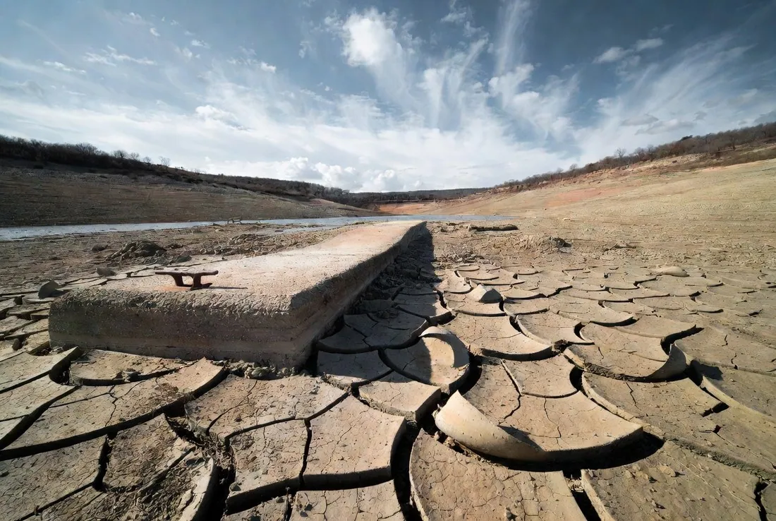 A dried cracked reservoir bed with a concrete boat ramp ending in open ground far short of the remaining water, a rusted mooring cleat still bolted in place
