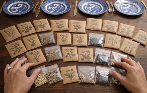 Hands sorting homemade heirloom seed packets and bags on a wooden dining table with Blue Willow china plates in the background