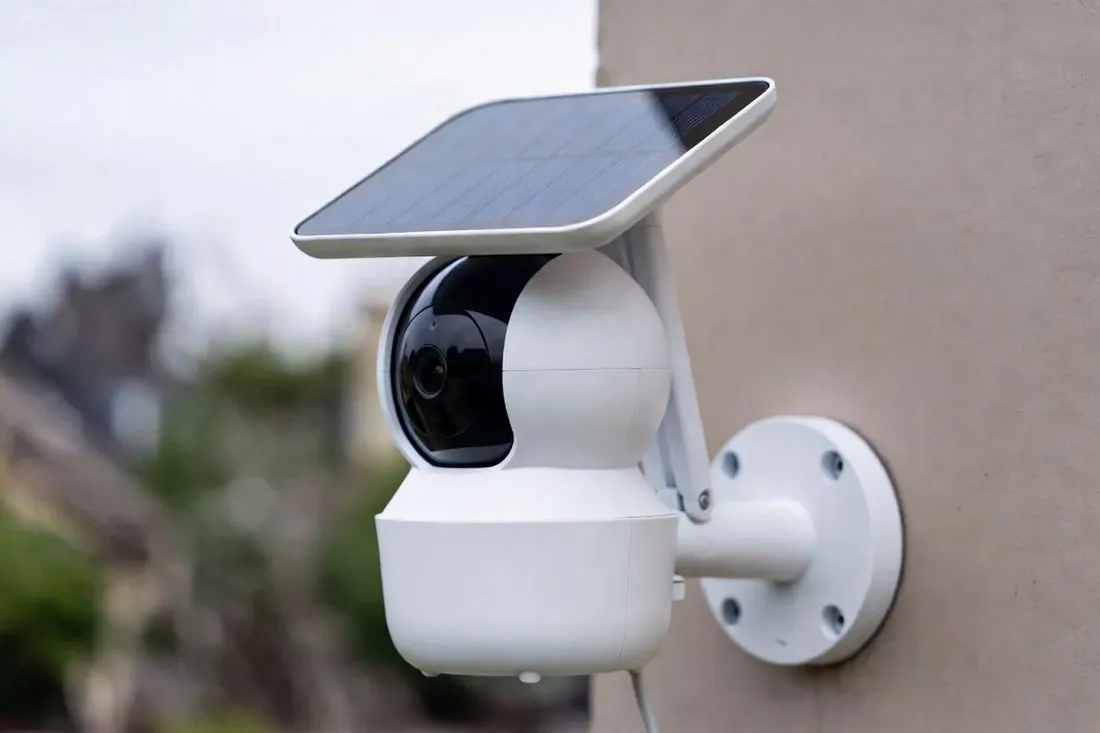 A white outdoor camera with a solar panel mounted above it, fixed to the wall of a house
