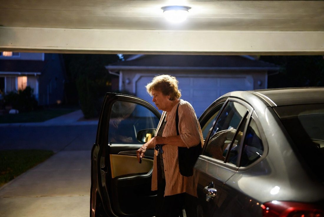 An older woman stepping out of her car in a lit driveway at night with her keys already in her hand and a bag on her shoulder