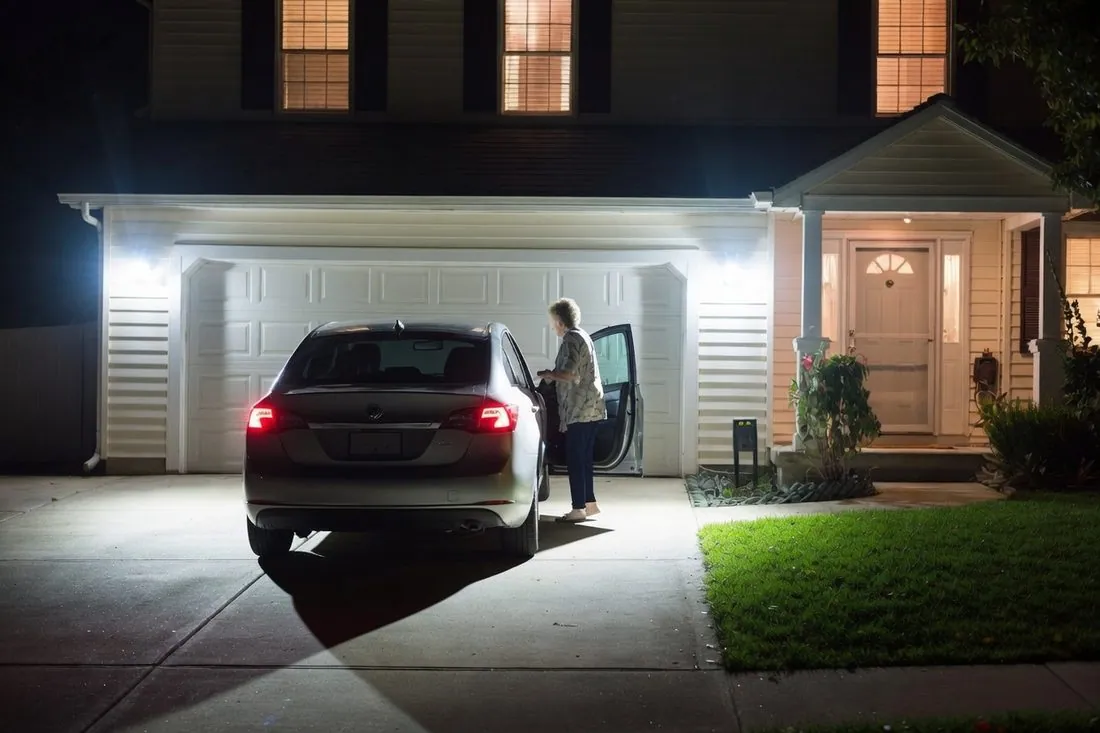 A car parked in a driveway at night with floodlights mounted above the garage door lighting the whole driveway and the ground beside the car, and an older woman getting out