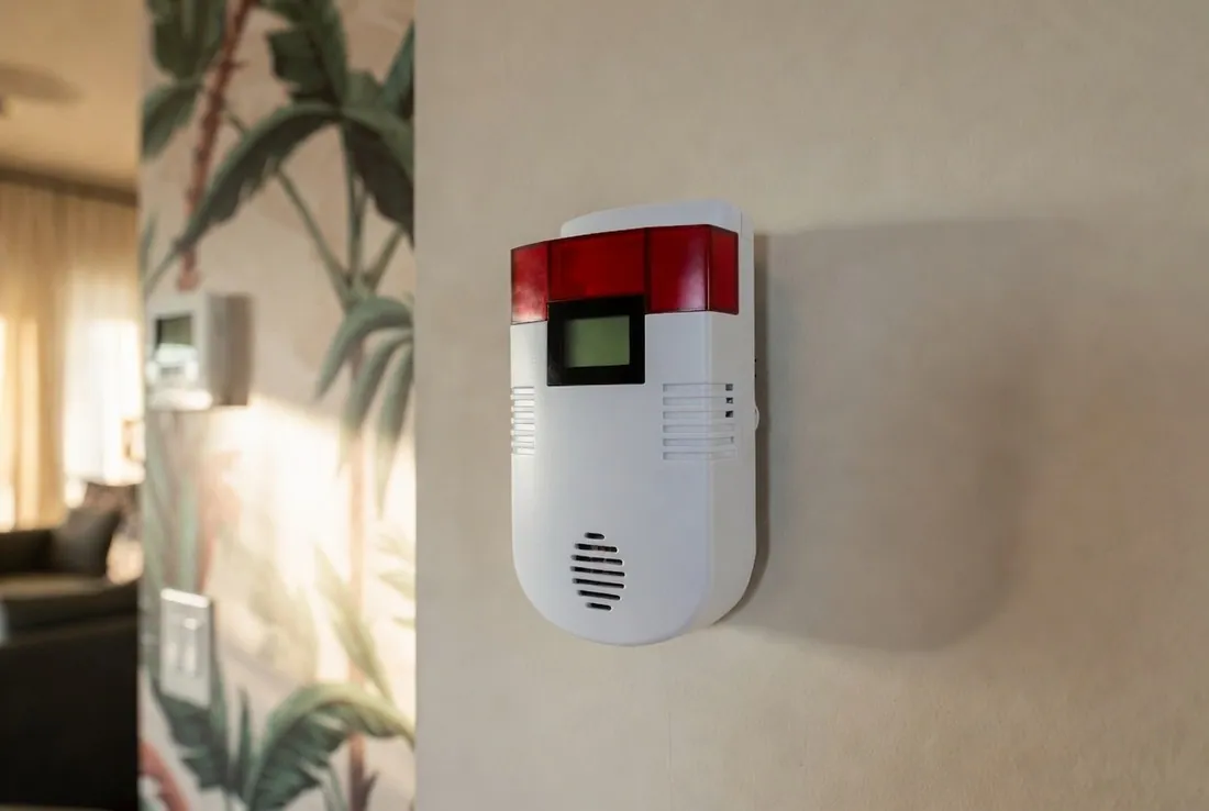 A white wall-mounted gas and carbon monoxide alarm with a red alert panel and a small display, fixed at about thermostat height on an interior wall