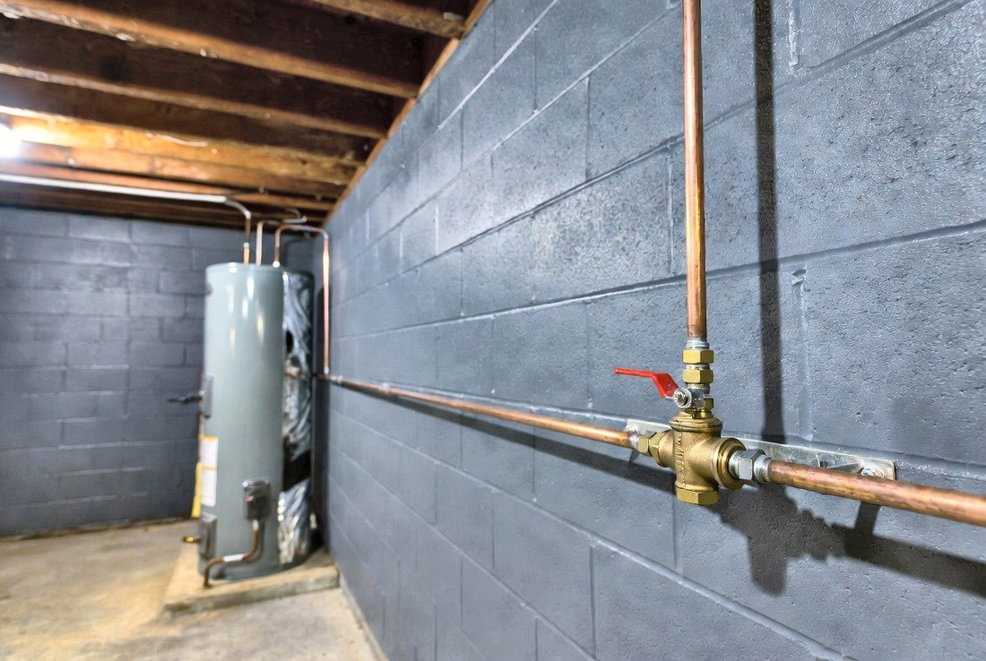 An interior main water shutoff on a basement wall: a horizontal copper line with a brass ball valve whose red lever is turned crosswise to the pipe, with a gas water heater against the wall behind it