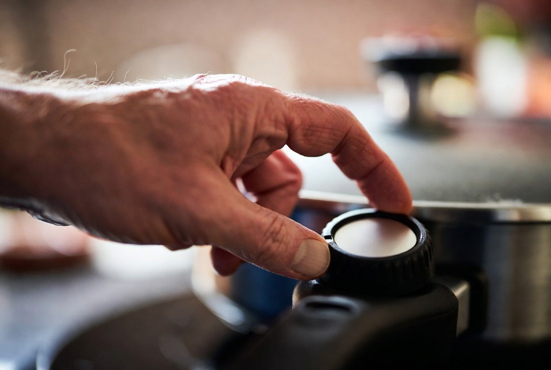 An older hand turning the temperature dial on the probe of an electric skillet, the dial face plain and unmarked, the pan soft in the background
