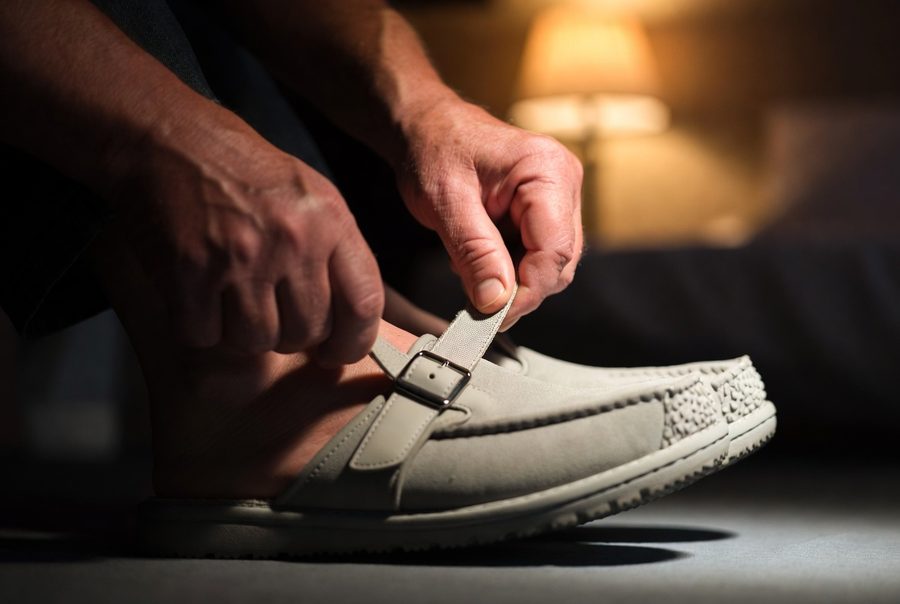 Older man's hands adjusting the buckle strap of a closed-toe house slipper with a treaded rubber sole, in warm evening light