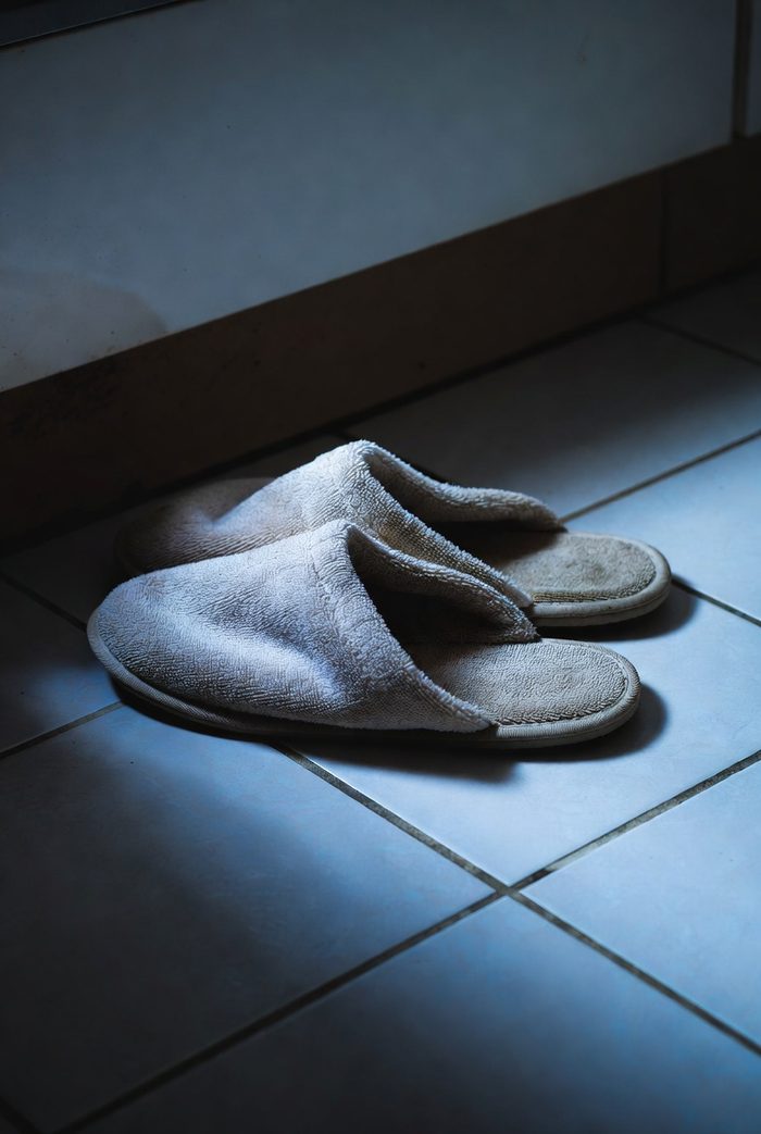 A worn-out pair of backless terry scuff slippers, flattened and stretched, sitting on smooth kitchen tile in cold light