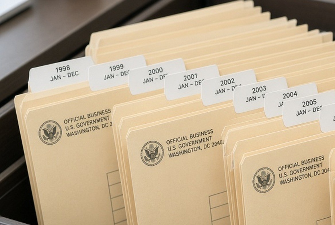 An open filing drawer with official envelopes filed upright in labelled folders, one for each year in sequence, a hand reaching for one of them