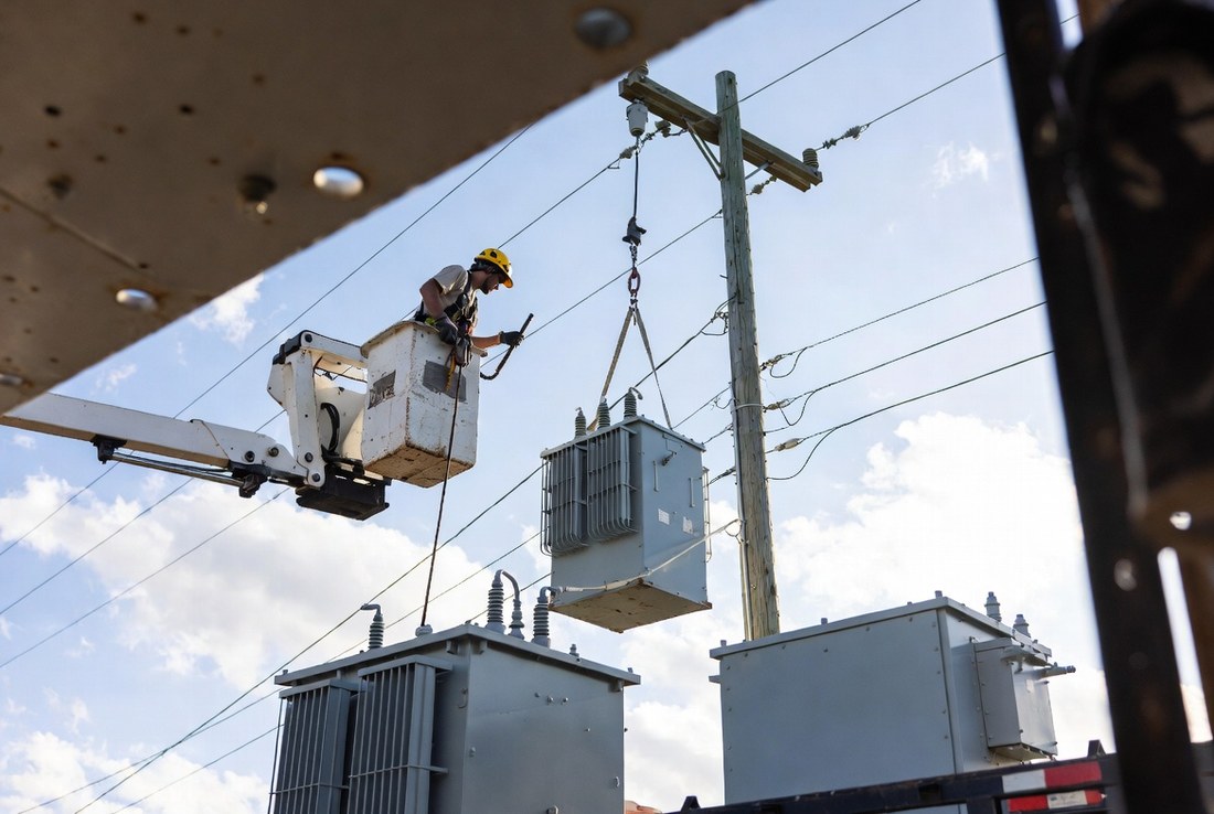A lineman in a bucket working at a pole while a replacement transformer hangs on a strap, with more transformers strapped to the flatbed below