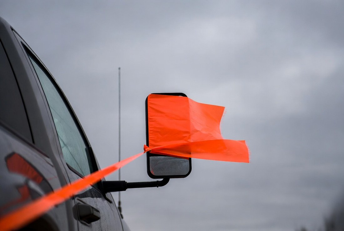 A length of bright orange flagging tape knotted to the side mirror of a pickup, streaming out straight in the wind against a flat grey winter sky