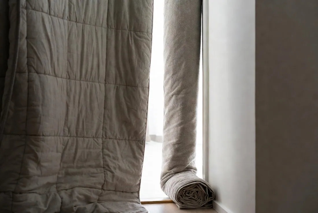 A heavy quilt hung across a doorway to seal a room, with a rolled towel laid along the bottom of the gap beside it