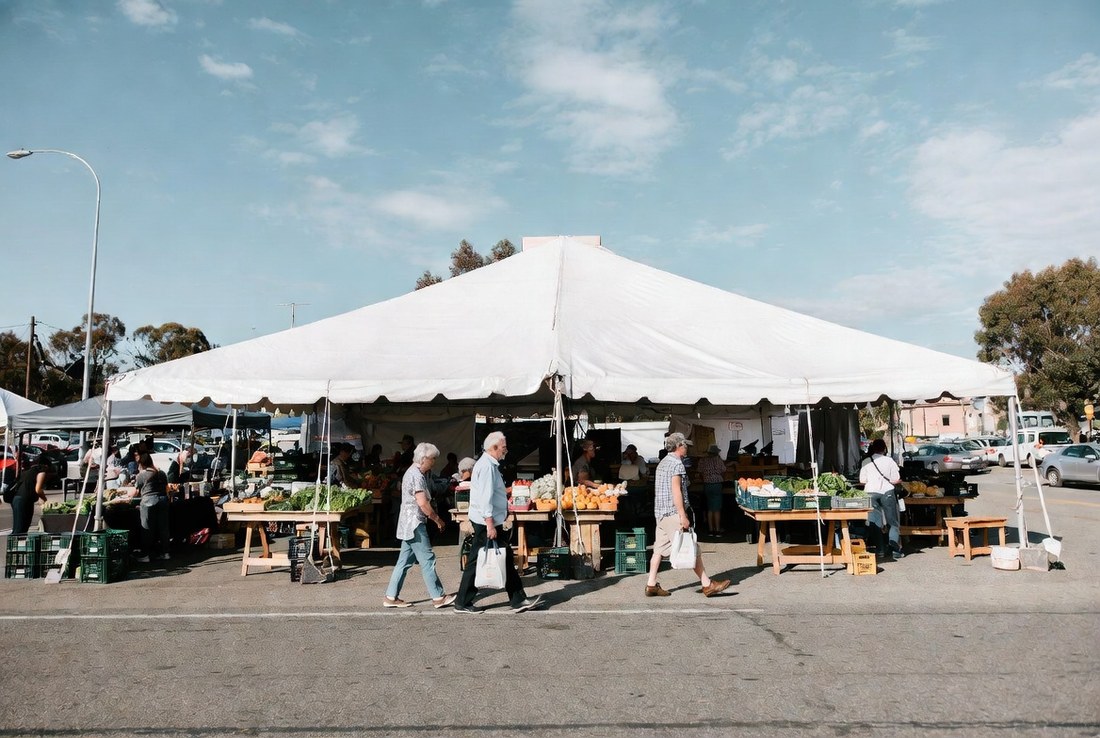 A farmers market under a large white tent on a weekday morning with a handful of older shoppers carrying bags and no crowd at all
